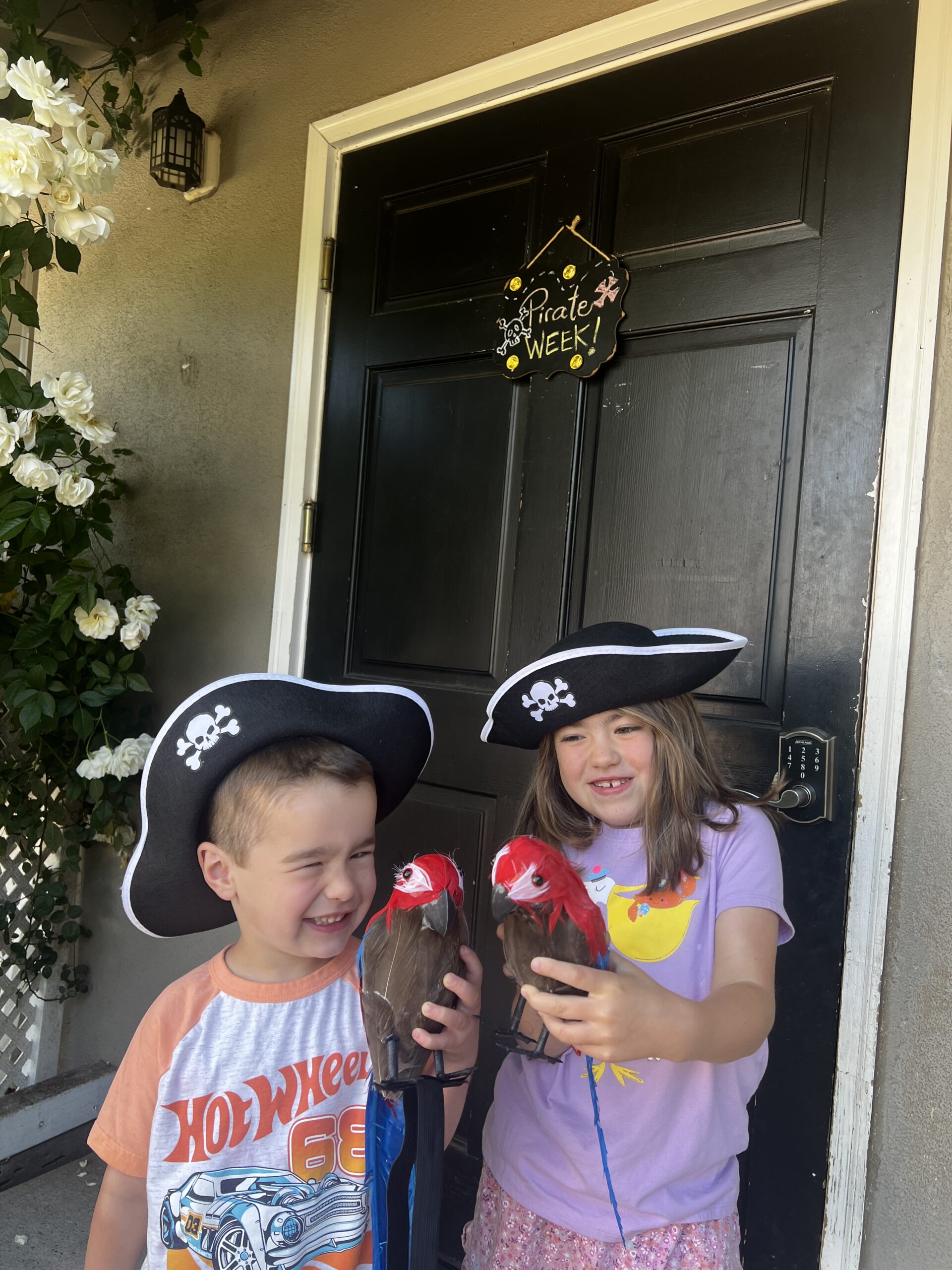 Two smiling children dressed as pirates holding toy parrots in front of a black door with a “Pirate Week!” sign.
