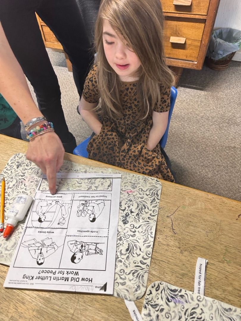 Child sitting at a classroom table while a teacher points to a worksheet about Martin Luther King Jr.’s contributions.