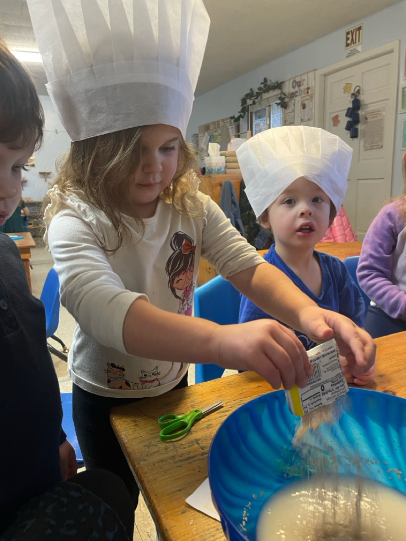 Preschool children wearing chef hats making bread from scratch by pouring yeast into a bowl.