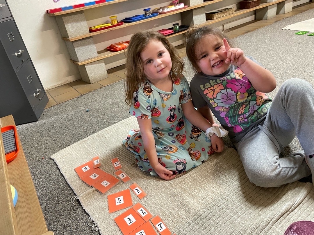Two young girls in a preschool classroom smiling while working on a word-matching Montessori activity with orange phonics cards.