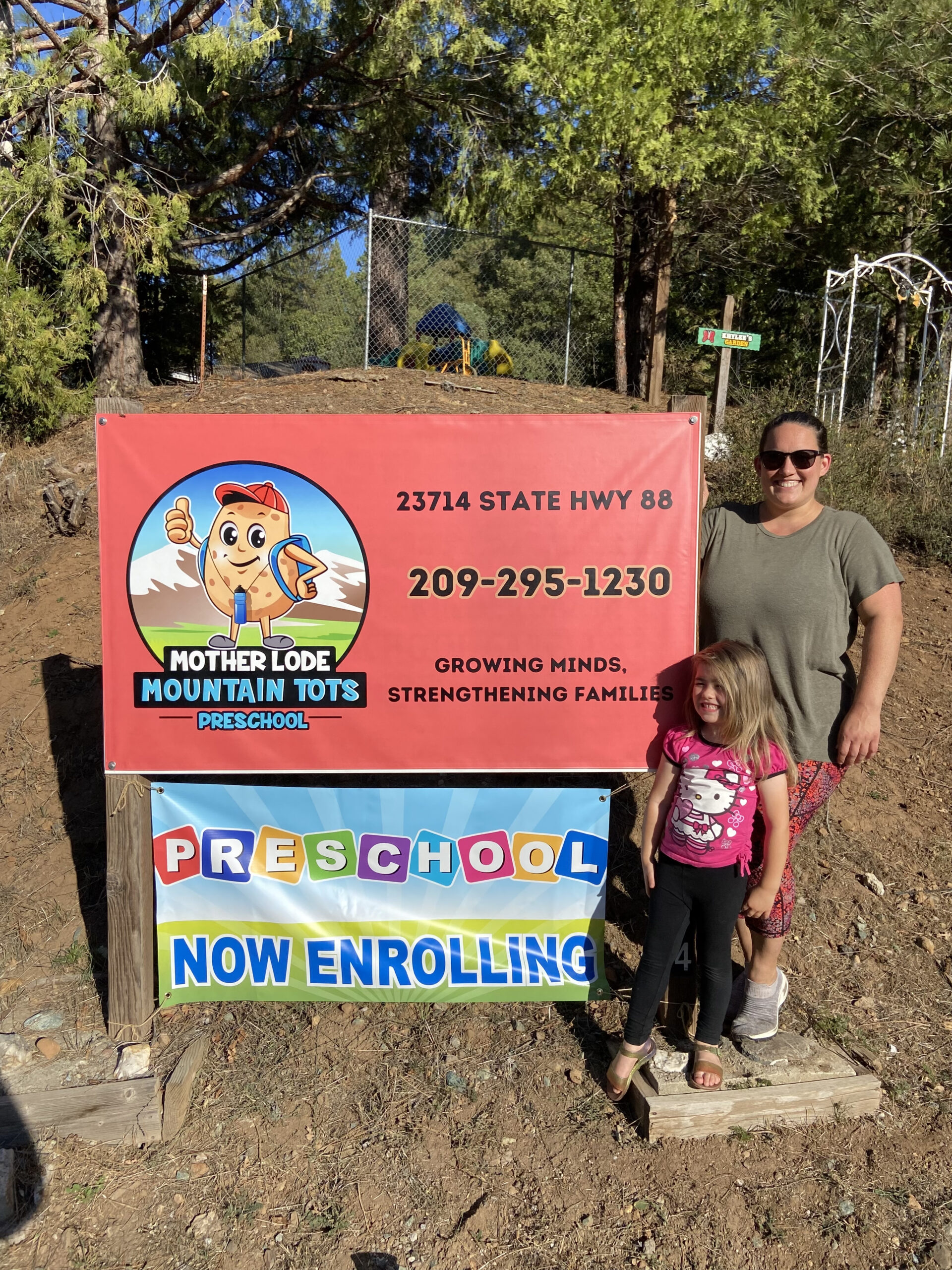 Ms. Erica and her daughter stand smiling next to the Mother Lode Mountain Tots enrollment sign outside the preschool in Pioneer, California.